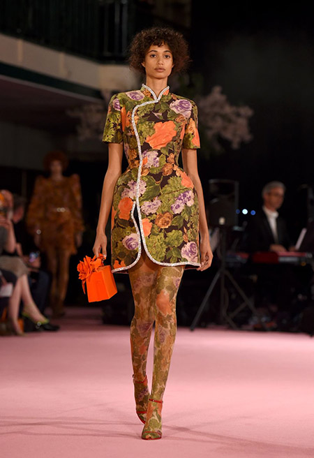 Desfile Richard Quinn na LFW Primavera / Verão 20, modelo usa vestido gola oriental e estampa floral em verde, lilás e laranja e meia calça na mesma estampa. Bolsa e sandália vermelhas.