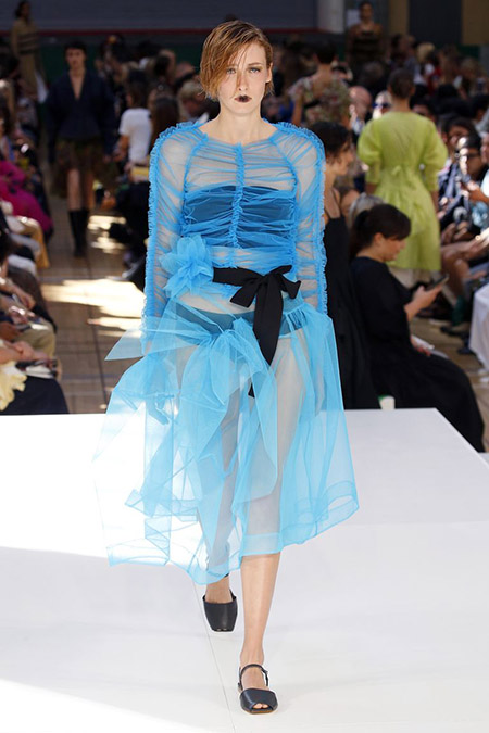 Desfile Molly Goddard na LFW Primavera / Verão 20, modelo veste vestido translúcido de tule azul, com top e calcinha pretas. Um laço preto amarra a cintura do vestido na frente.