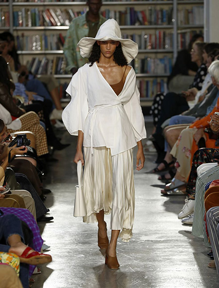 Desfile Rejina Pyo na LFW Primavera / Verão 20, modelo usa ampla blusa off white com amarração trespassada na frente. Saia bege, sapatos marrons e chapéu mole de ponta branco.