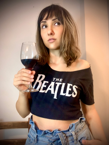 Natalia está com franja, segura uma taça de vinho em uma das mãos e veste uma camiseta curta dos Beatles.