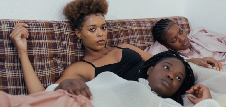Mulheres pretas deitadas em um sofá, algumas olham para a lente e te convidam a pensar sobre racismo na nossa sociedade e em cada um de nós.