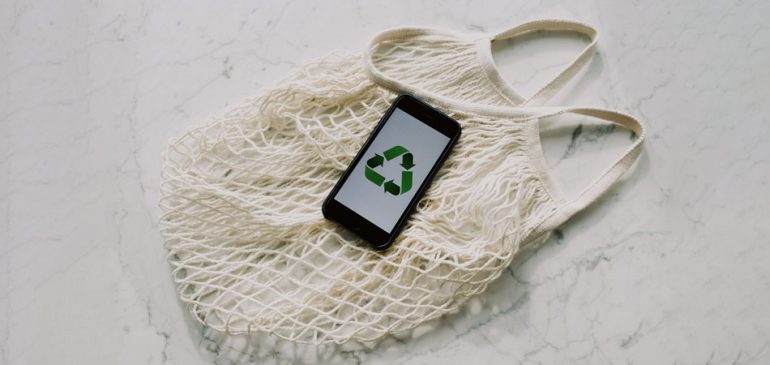 Celular com logo de reciclagem na tela (3 setas que formam um triângulo), sobre uma sacola branca de linha. A ideia é falar sobre sustentabilidade, tendências da moda e como o guarda-roupa compartilhado pode ajudar.